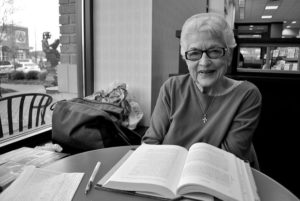 a white-haired older white woman sits smiling at a table with a book in front of her. 